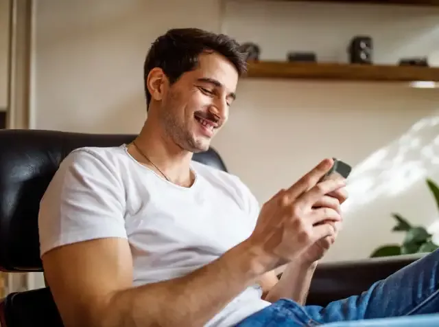 Ein Mann sitzt auf einem Sofa und blickt begeistert auf sein neues Smartphone, das er von seinem Arbeitgeber über den Pluxee Benefit Elektronik Geschenke bekommen hat.