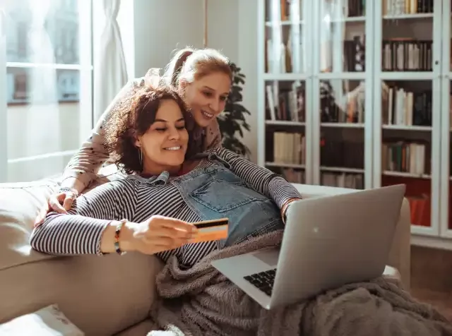 Auf dem Sofa sitzen zwei Frauen und erledigen ihre Online Einkäufe mit einer Pluxee Prepaidkarte. Dank der Mitarbeiterbenefits von Pluxee erhalten sie eine Internet Pauschale von ihren Arbeitgebern.