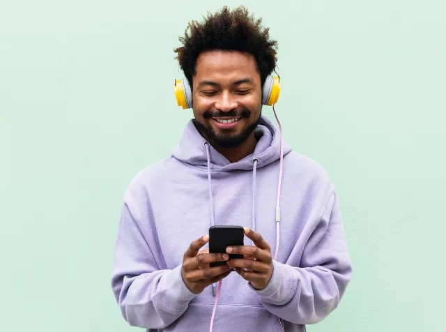 in Mann mit Smartphone in der Hand, Kopfhörern und lila Kapuzenpulli, hört Musik und lächelt, weil er von seinem Arbeitgeber den Pluxee Benefit Mobilfunk Zuschuss erhält.